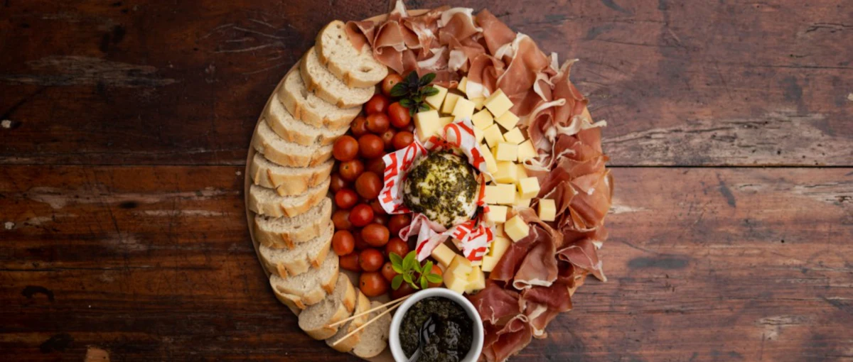 A round charcuterie board with bread, cheese, tomatoes and olives on dark wood